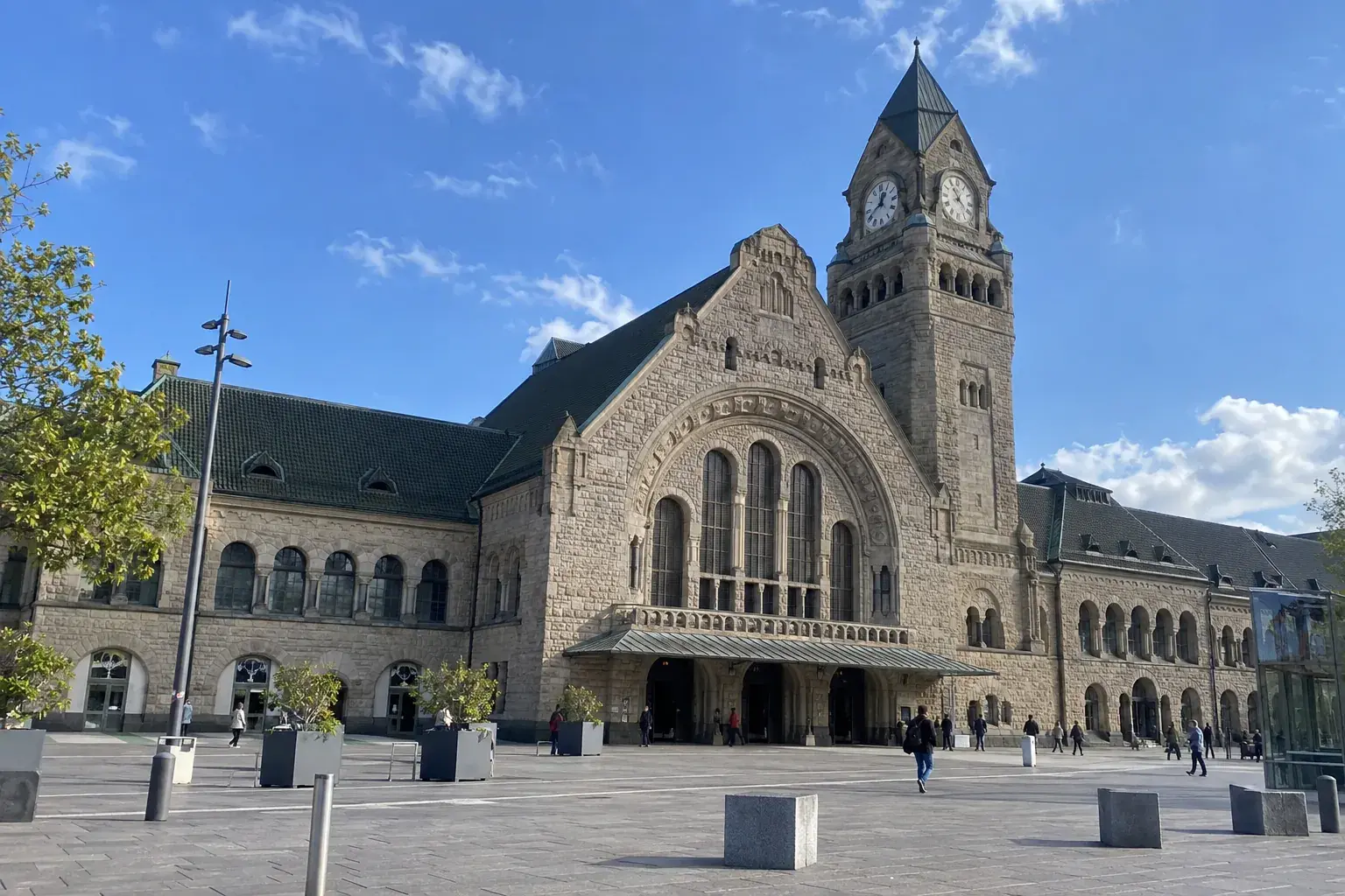 La façade de la gare de Metz-Ville, long bâtiment néo-roman en grès clair avec sa tour de l’horloge, et la place devant la gare sous un ciel bleu