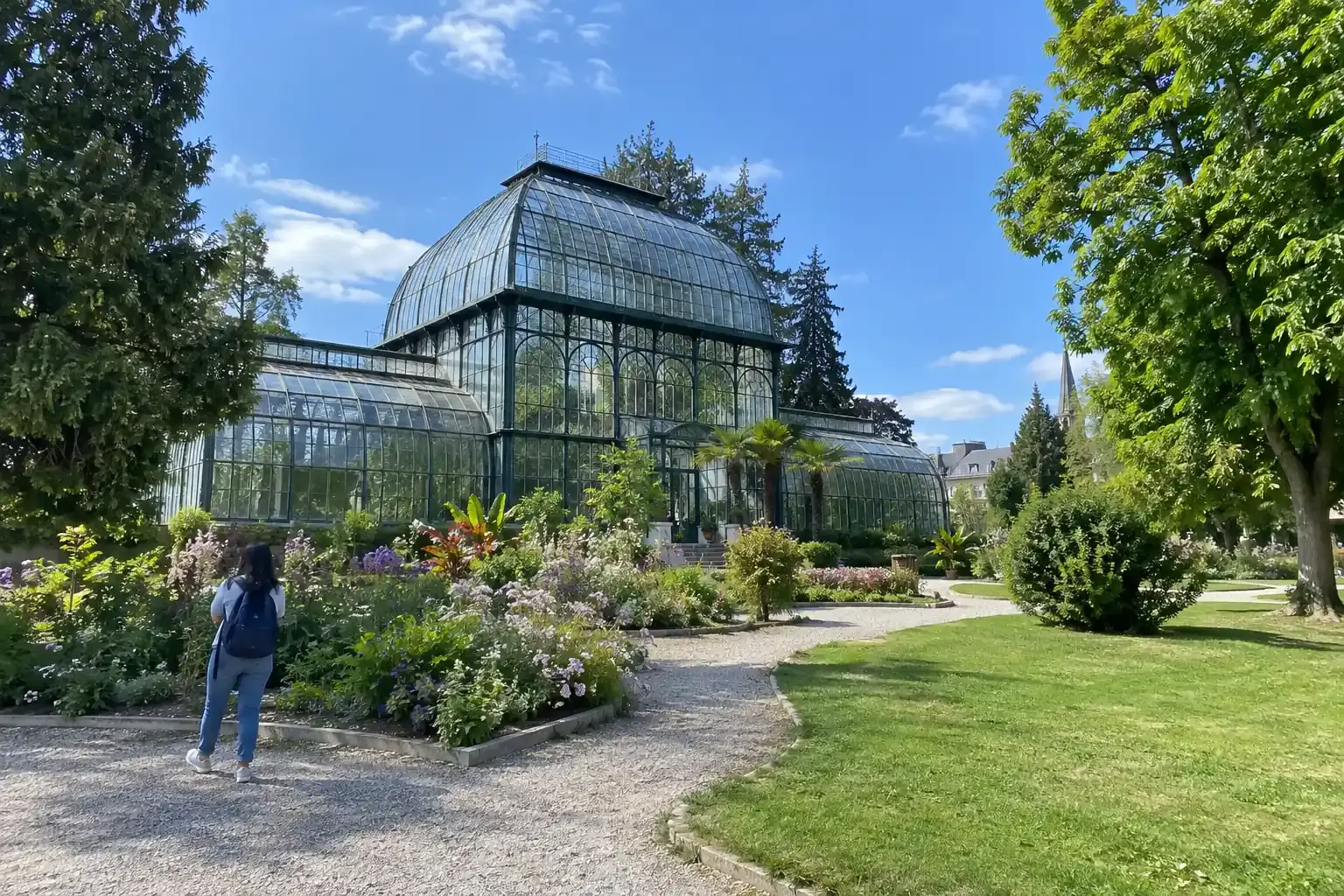 Une serre ancienne en verre et métal dans un jardin botanique, allées de gravier, massifs fleuris, pelouse et grands arbres sous un ciel bleu