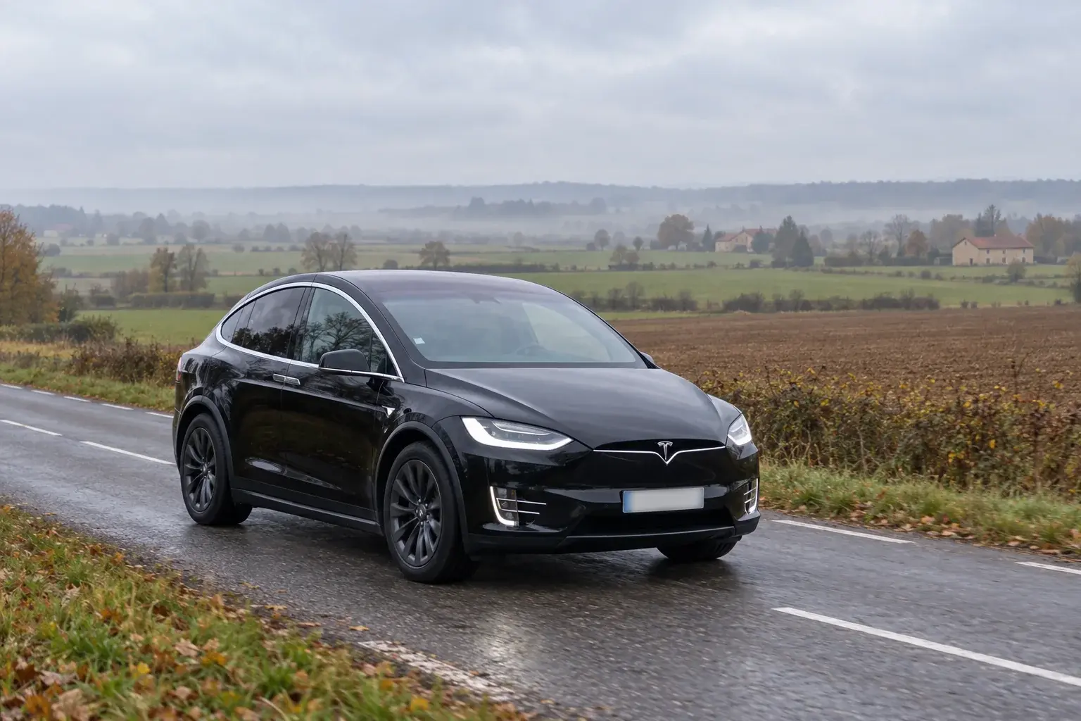 Une Tesla Model X noire sur une petite route de campagne qui traverse un plateau agricole de Lorraine, entre des labours et des prés, par un matin d’automne couvert et brumeux