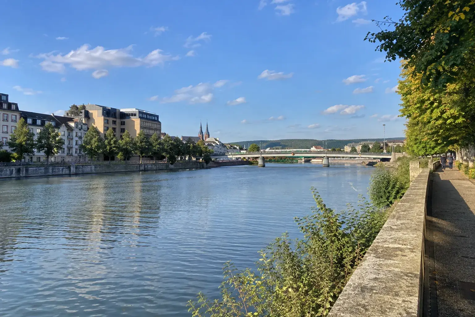 Les quais de la Moselle à Thionville : le fleuve large et calme, un pont, les immeubles de la ville et des arbres sur la berge, sous un ciel bleu