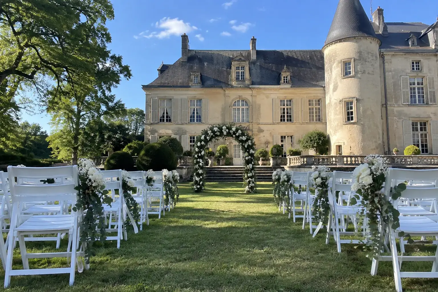 Une allée de chaises blanches ornées de fleurs blanches et de verdure dans le parc d’un château en pierre claire, préparée pour une cérémonie de mariage