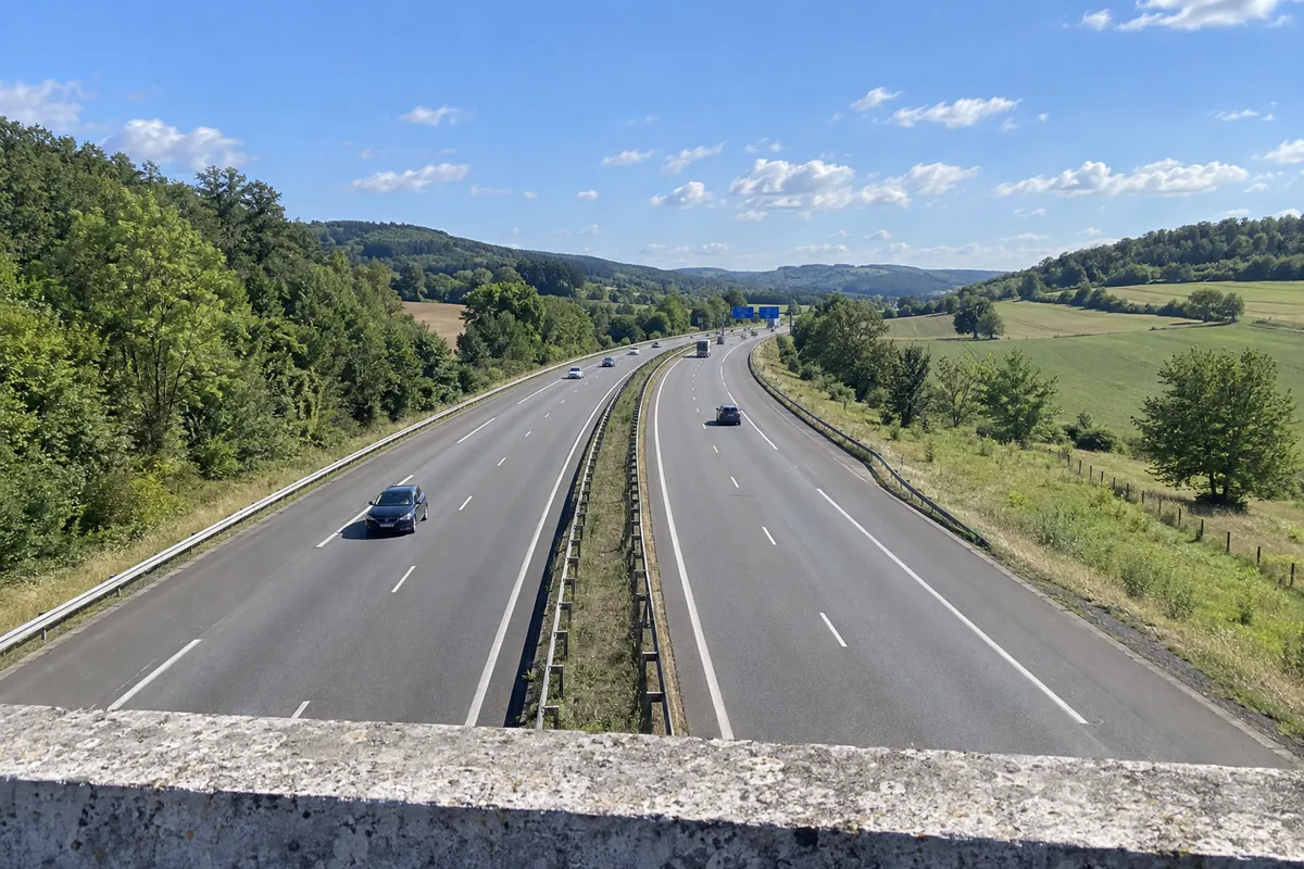 Une autoroute à deux fois deux voies entre des collines boisées et des champs, vue depuis un pont par une matinée claire